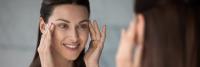 Smiling woman examines skin around eyes by looking in a mirror.